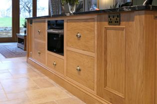 Wooden kitchen cabinets with brass handles and a dark countertop are in focus. In the background, green cabinets add a contrasting touch. Warm ambiance. 