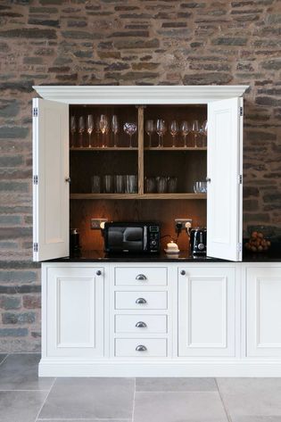 White kitchen cabinet against a brick wall, open to reveal glassware on shelves. Below are a black microwave, a coffee machine, and a toaster.