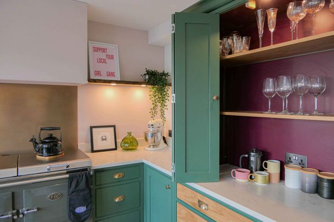 A shaker kitchen with green cabinets, open shelf with glasses, mugs, and a kettle on a stove. Sign reads "Support Your Local Girl Gang." Bright, welcoming vibe.