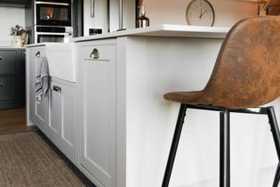 Shaker style kitchen with white island, farmhouse sink, and brown leather stool. Metal appliances and a wall clock create a sleek, contemporary vibe.