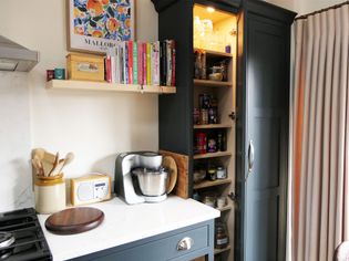 Shaker kitchen corner with dark cabinets. Open pantry reveals jars and cans. Countertop holds a mixer, radio, and utensil jar. Shelves with cookbooks. Cozy ambiance.