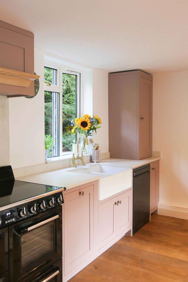 Bright pink shaker kitchen with a white worktop and brass tap. A vase of vibrant sunflowers sits on the windowsill, adding a cheerful touch.