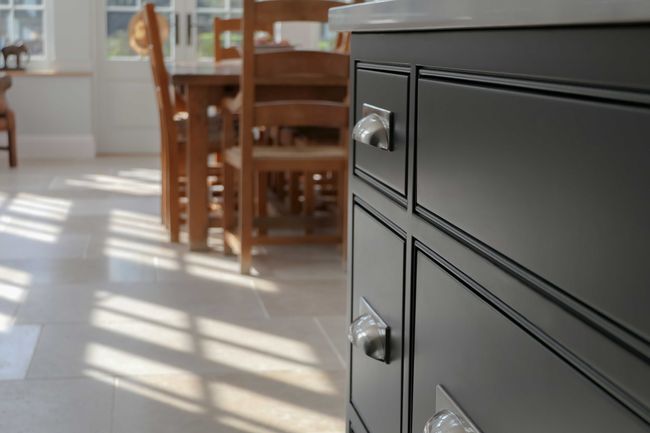Sunlight streams through a window, casting shadows on a tiled floor. A dark grey cabinet with metallic handles is in focus, with wooden chairs in the background.