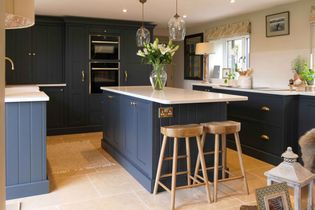 Shaker kitchen with a dark blue island, wooden stools, and white worktop. Pendant lights hang above, and flowers in a vase add a fresh touch.