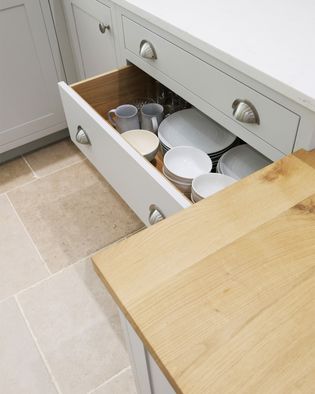 Open kitchen drawer with neatly stacked white plates, mugs, and glasses against light cabinetry. Natural wood and stone-tiled floor evoke warmth.