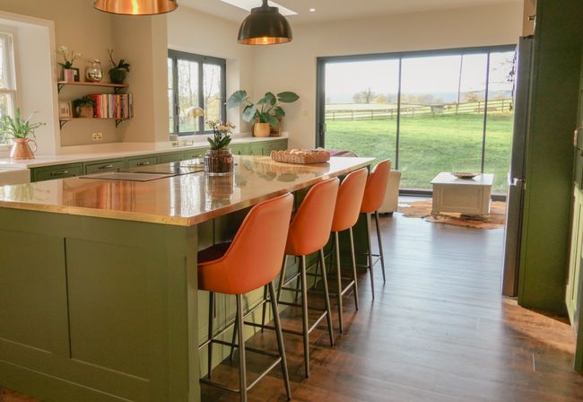 A large island with a brass worktop, pendant lighting above and four orange leather bar stools with a countryside backdrop out of the large windows.