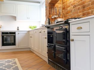 Shaker kitchen with light cabinets, a dark vintage-style stove, and brick wall. Bright, clean atmosphere with utensils hanging neatly.