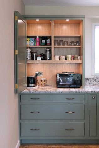 Open shaker kitchen cabinet with green doors, displaying coffee supplies, mugs, a kettle, and a microwave on a marble countertop. Bright, organized space.