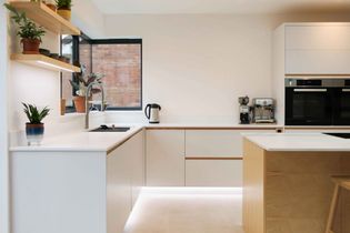 Modern kitchen with sleek white cabinets and wood accents. Features a built-in oven, coffee maker, and potted plants on shelves for a fresh feel.