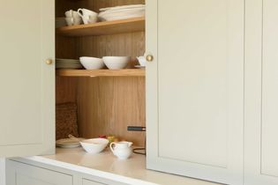 Open kitchen cabinet with light shelves holding white bowls, plates, and cups. A tray and condiments are on the countertop, conveying an organized, cozy kitchen feel.