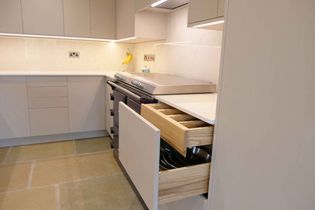 Modern kitchen with sleek light grey cabinets, open cutlery drawer, and under-cabinet lighting. Stove on the left.
