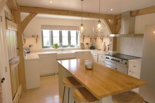 Shaker style kitchen with wooden beams, featuring a central island with wooden top and stools. Bright light from large window and two pendant lights over the island.
