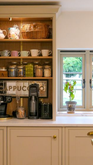 Cream Shaker kitchen cabinetry with open wooden shelving, crockery and jars, a countertop, and a bright window with a potted plant.
