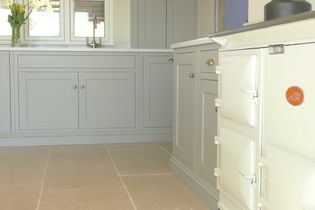 Bright kitchen with cream cabinets, an off-white AGA stove, and beige tiled floor. A vase of yellow tulips sits on the counter by a window.