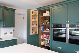A shaker kitchen with green cabinets and a pantry door open, revealing spices and various groceries. Stainless steel double ovens are built into the cabinetry.