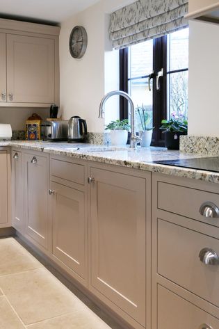 Shaker kitchen with beige cabinets, worktops, and a stainless steel sink. Bright window with potted plants and a wall clock enhance the elegant ambiance.