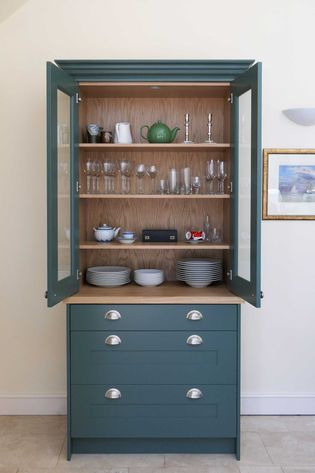 Green cabinet with open glass doors reveals neatly arranged tableware, including glasses, a green teapot, and candlesticks, creating an organized and elegant look.