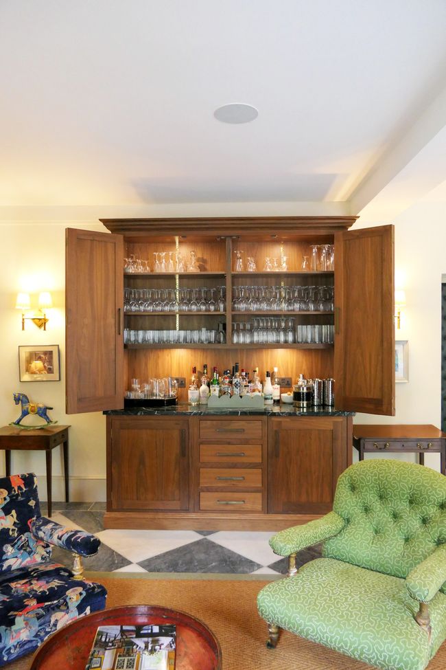 A well-decorated living space featuring a wooden walnut bar with open shelves displaying glassware and various bottles, accompanied by stylish furniture including a green upholstered chair and a patterned sofa.