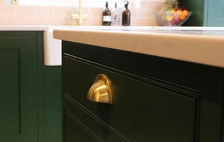 Close-up of a kitchen with dark green cabinets, a shiny brass handle, a light countertop, and soap dispensers. The tone is modern and elegant.
