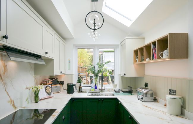 Green and cream cabinetry in a u-shaped layout with a kitchen sink infront of the window. Different appliances out on the marble effect worktop.