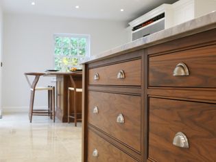 Warm, shaker kitchen with wooden island, sleek drawers, metallic handles. Soft lighting, neutral tiles, and window enhance the inviting atmosphere.
