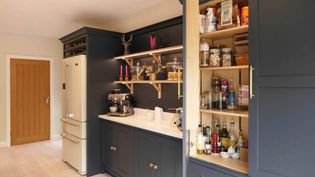 Shaker kitchen with dark grey cabinets, light countertop, a white fridge, and open shelves displaying jars and decor. Open pantry shows condiments and bottles.