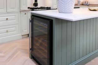 Shaker kitchen with light green cabinetry featuring gold knobs, an inset wine fridge, and a white countertop. The tone is elegant and fresh.