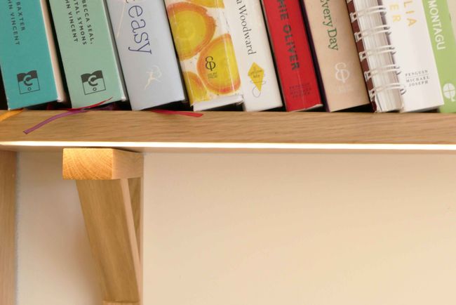 Books are neatly lined on a wooden shelf with colourful covers and bookmarks against a cream wall. The scene feels organised and inviting.