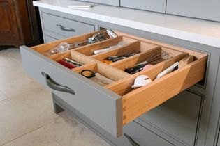 Open kitchen drawer with organized wooden dividers, holding utensils like spoons, scissors, and peelers. Grey cabinetry and quartz worktop visible.