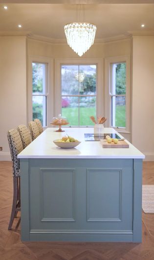 A Shaker kitchen with a light blue island, three high-backed chairs, a large chandelier, and a view of a garden through a bay window.