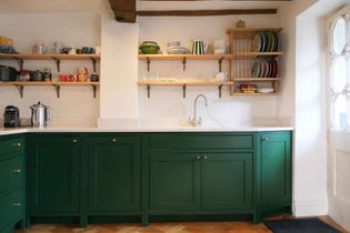 A cosy kitchen with deep green cabinets and white worktops. Two wooden shelves hold colorful dishes and mugs. A chrome taps is centered above the sink.