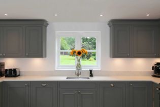 A shaker kitchen features grey cabinets, a light countertop, and a centered window with a vase of sunflowers. The scene feels bright and welcoming.