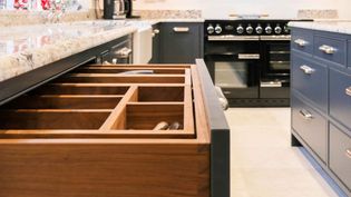 Open kitchen drawer with wooden utensil organiser, surrounded by granite countertop, dark cabinetry, and a sleek black oven.