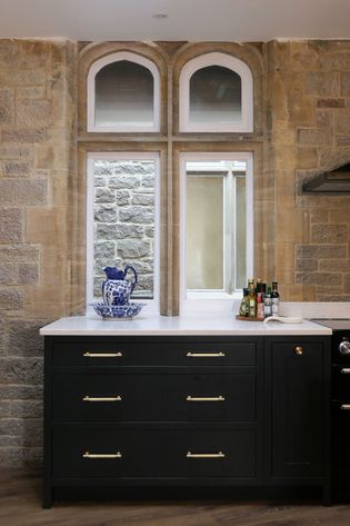 A kitchen interior features rustic stone walls and arched windows. A dark grey cabinet with gold handles sits below, topped with a blue vase and condiments.