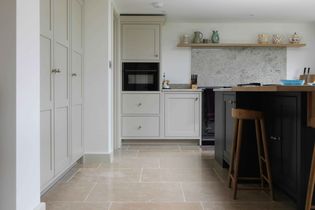 Shaker style kitchen with light grey cabinets, black oven, and blue centre island with wooden stools. Stone backsplash, countertop accessories, calm ambiance.