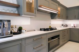Shaker kitchen with sleek grey cabinets, gold handles, under-cabinet lighting, and white countertops. Features a coffee machine and a gas stove.