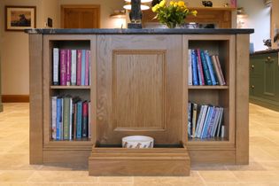 Wooden kitchen island with black countertop, featuring bookshelves on either side filled with colourful books. A drawer below opens to reveal a dog bowl.