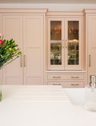 Pink flowers on the left, pink shaker cabinetry with a white worktop. Glassware cupboard in the background.