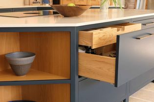 Shaker kitchen island with a sleek white countertop and navy-blue cabinets. Open drawers reveal wooden interiors. A ceramic vase and a fruit bowl add warmth.
