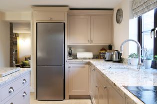 Shaker kitchen with beige cabinets, stainless steel fridge, and speckled worktops. Bright light, cozy feel, with a window plant and toaster.