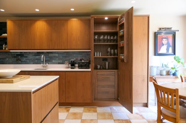 Modern walnut kitchen with a white island, dark tiled backsplash, sink, coffee machines, and a potted plant.