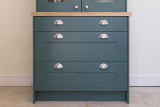 A green cabinet with three drawers, featuring silver cup handles and a wooden countertop. The cabinet stands against a light wall on a tile floor.