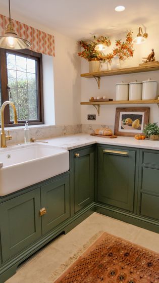 A cosy kitchen featuring a farmhouse sink, green cabinets, wooden shelves with decorative items, and warm lighting.