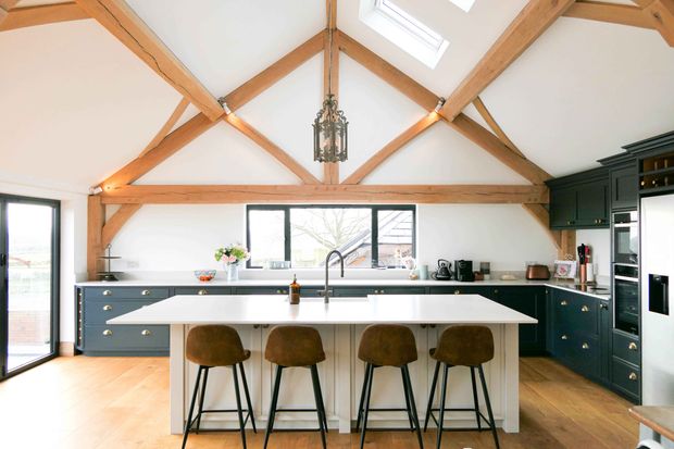 Shaker kitchen with exposed timber beams, dark cabinetry, a large white island, four bar stools, and a central hanging chandelier.