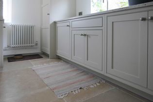 Bright kitchen with light cabinets and a striped rug on a tiled floor. A window on the right, radiator on the left, and a dog's bowl in the corner.