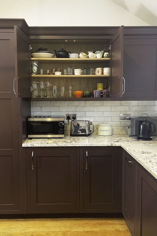 A kitchen with dark red cabinets and granite countertops, featuring a microwave, kettle, and various dishes on an open shelf.