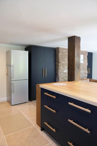 Modern navy blue kitchen with floor to ceiling cabinets, stone wall in the background, large free standing fridge-freezer to the left. Kitchen island and wooden beam to the right.