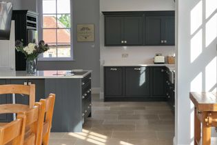 Bright shaker kitchen with dark cabinetry, light countertops, and beige tile flooring. Wooden chairs and sunlight through windows, creating a cozy ambiance.