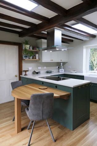 Shaker kitchen with exposed beams, green cabinets, and white quartz. A wooden table extends from the island, paired with gray chairs. Creating a cosy ambiance.