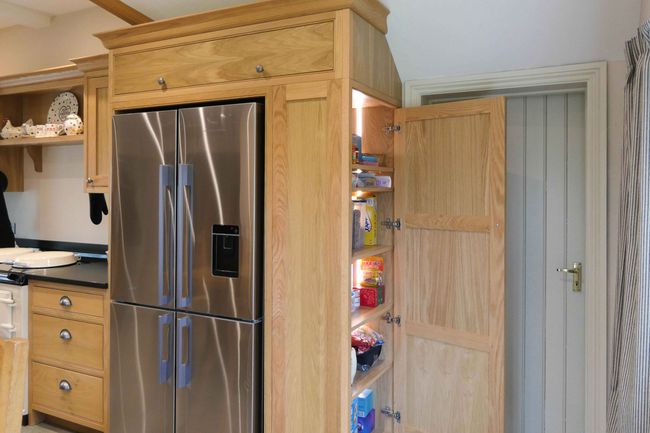 Wooden kitchen with a double-door stainless steel fridge. Adjacent open pantry reveals snacks and groceries, creating a welcoming and organized space.